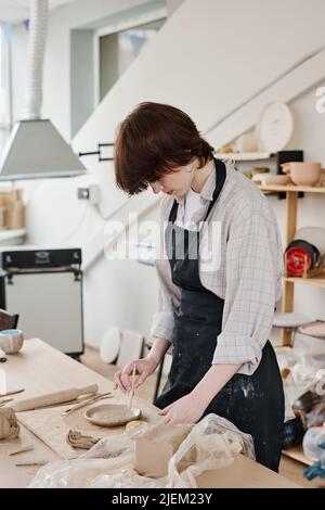 Jeune femme en vêtements de travail coupant l'ornement sur plaque d'argile brute tout en se pliant sur la table avec des fournitures pour créer de la faïence Banque D'Images