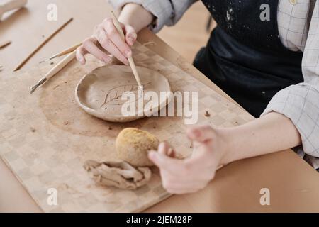 Mains de jeunes femmes maîtres en poterie coupant la feuille sur la pièce en plaque d'argile à bord tout en étant assis à table dans l'atelier Banque D'Images