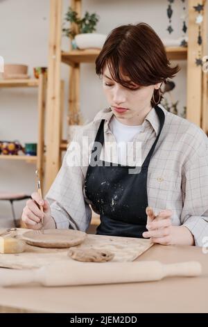 Brunette femelle dans un tablier assis sur le lieu de travail et utilisant une pince à main en bois pour couper des ornements sur la surface des articles en argile crue Banque D'Images