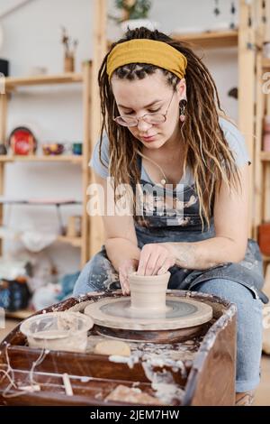 Jeune femme créative en vêtements de travail assise devant la roue de poterie rotative dans l'atelier et faisant de nouveaux articles en faïence à vendre Banque D'Images