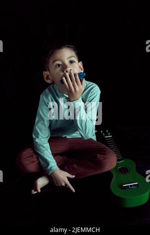 garçon latino-américain assis sur le sol jouant l'harmonica et avec un ukulele sur le sol à côté de lui. fond noir. concept de l'éducation musicale Banque D'Images