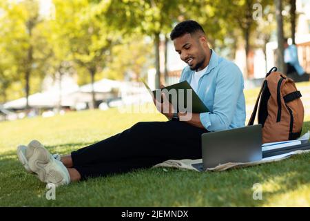 Concept de Studentship. Un homme afro-américain heureux qui esquoit ou prend des notes, fait ses devoirs tout en étant assis à l'extérieur Banque D'Images