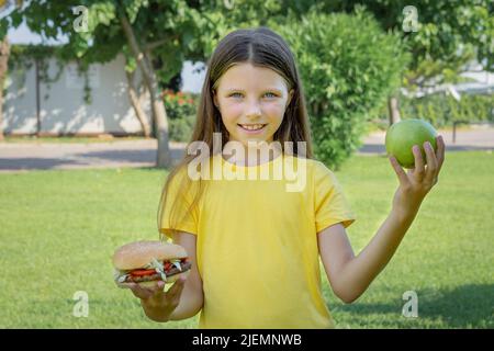 Une adolescente choisit entre un hamburger et une pomme en plein air dans le parc. Banque D'Images