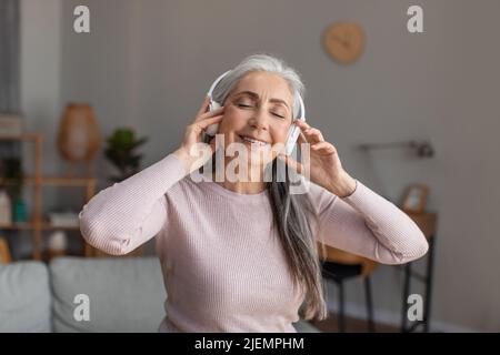 Souriante, jolie femme européenne mûre avec cheveux gris dans un casque sans fil, vous permet de profiter de votre musique préférée et de votre temps libre Banque D'Images