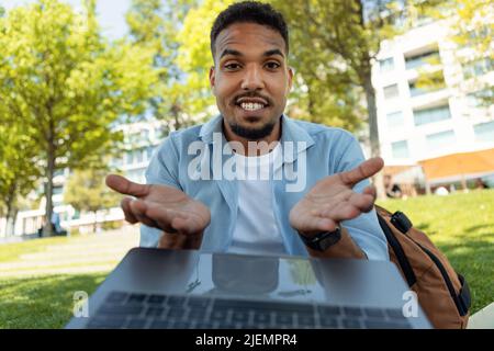 Jeune homme noir enthousiaste parlant à la famille ou à un ami sur un ordinateur portable, faisant des gestes à la webcam, assis à l'extérieur Banque D'Images
