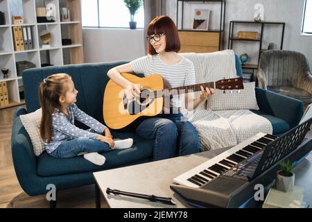 Maman et petite fille heureuse en musicothérapie en jouant de la guitare dans la salle de musique. Enseignant aidant la jeune femme élève dans la leçon de guitare. Détente à la maison. Banque D'Images