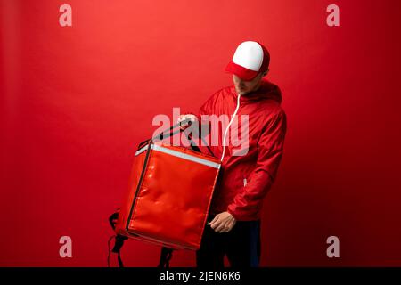 Professionnel de livraison employé homme 20s dans Red Cap T-shirt uniforme de travail travailler comme le concessionnaire de messagerie avec sac thermique rouge sac à dos isolé sur rouge Banque D'Images