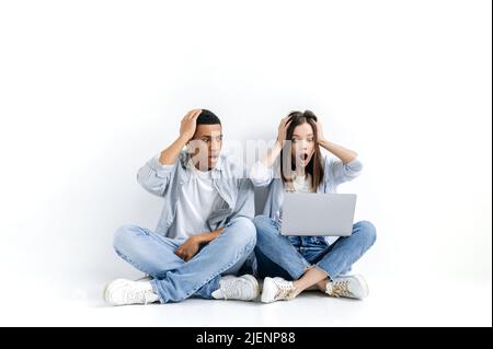 Un jeune homme hispanique et une femme caucasienne choqués, élégamment habillés, assis au sol sur un fond blanc isolé avec un ordinateur portable, regardant un écran dans la surprise, ont vu des nouvelles inattendues Banque D'Images