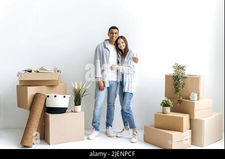 Heureux moderne multiracial couple dans l'amour, emménagé dans leur nouvelle maison ou appartement loué, debout contre le mur blanc, embrassant entre des boîtes en carton d'articles ménagers, regarder l'appareil photo, sourire Banque D'Images