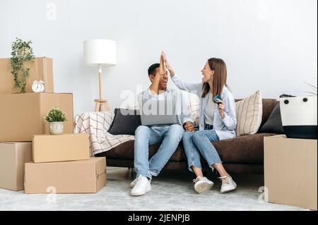 Des jeunes mariés heureux de différentes nationalités, un type hispanique et une fille caucasienne, s'assoient dans leur nouvel appartement dans le salon, parmi des boîtes en carton avec des choses, font des achats en ligne pour la maison en utilisant un ordinateur portable Banque D'Images