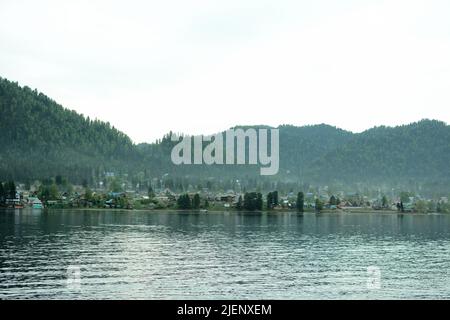 Vue panoramique sur le lac jusqu'à un petit village sur les pentes d'une montagne couverte de brouillard léger. Village d'Iogach, Altaï, Sibérie, Russie. Banque D'Images