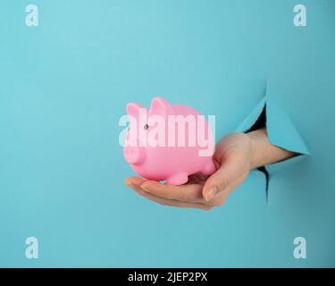 Une main femelle qui dépasse d'un trou d'un fond bleu tient une tirelire rose. Banque D'Images