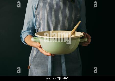 Dans un studio moderne, faire cuire une femme anonyme dans un bol de support à tablier avec une spatule tout en préparant la cuisson sur fond noir Banque D'Images