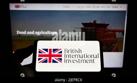 Personne tenant un téléphone portable avec le logo de British International Investment (BII) sur l'écran en face de la page Web d'affaires. Mise au point sur l'affichage du téléphone. Banque D'Images