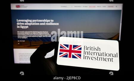 Personne tenant un téléphone portable avec le logo de British International Investment (BII) sur l'écran en face de la page Web d'affaires. Mise au point sur l'affichage du téléphone. Banque D'Images