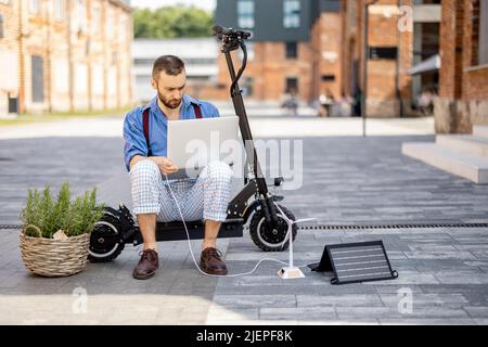 L'homme charge l'ordinateur portable avec des panneaux solaires tout en étant assis sur un scooter électrique à l'extérieur Banque D'Images