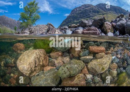 Rochers et cailloux sous la rivière COE lors d'une journée écossaise ensoleillée. Les trois montagnes sœurs, le ciel bleu et un arbre sont tous visibles au-dessus de la ligne d'eau Banque D'Images