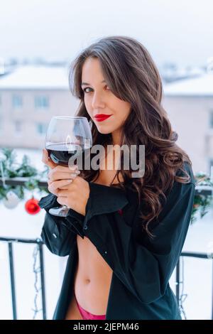 Portrait d'une jeune femme souriante avec de longs cheveux foncés ondulés, maquillage portant une chemise noire non boutonnée, sous-pantalons rouges, debout sur le balcon, tenant le verre W Banque D'Images