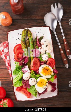 Asperges, tomates, laitue, mozzarella, sésame noir, lin, huile d'olive salade et œuf dur, sur plaque rectangulaire en céramique sur une table en bois sombre b Banque D'Images