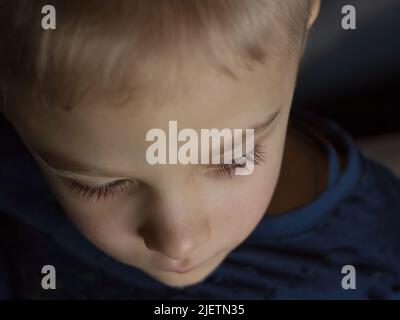 Le visage d'un garçon triste et clair avec des cils longs foncés pris d'en haut dans une clé sombre Banque D'Images