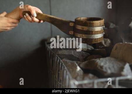 Main de femme tenant une louche en bois et versant de l'eau sur des pierres chaudes dans le sauna Banque D'Images