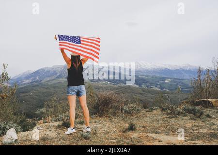 Jeune femme debout sur une falaise et agitant le drapeau américain tout en regardant les montagnes en arrière-plan. Une fille qui agite un drapeau américain debout Banque D'Images