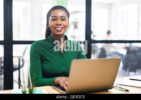 Jeune femme d'affaires afro-américaine accueillante et accueillante utilisant un ordinateur portable dans un bureau contemporain sur le lieu de travail, jeune femme d'affaires multiraciale confiante regardant l'appareil photo et souriant Banque D'Images
