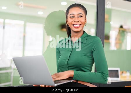 Femme d'affaires afro-américaine avec style décontracté se tient dans un espace de travail moderne et tient un ordinateur portable, une femme freelance ou une étudiante avec un ordinateur portable à l'intérieur et regarde avec joie Banque D'Images