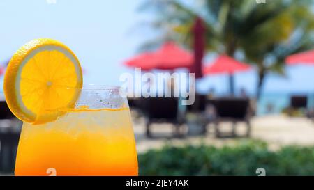 Boisson à l'arrière-plan avec une tranche d'orange au bar de la plage, au bord de la piscine, avec mer, palmiers et chaises longues en arrière-plan. Rafraîchissement boisson froide. Concept Banque D'Images