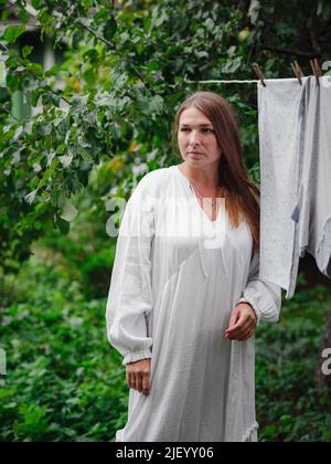 femme caucasienne d'âge moyen en robe blanche faisant des devoirs, pendant des vêtements sur la corde à linge dans la rue dans la cour de la maison de village, concept de jour de blanchisserie Banque D'Images