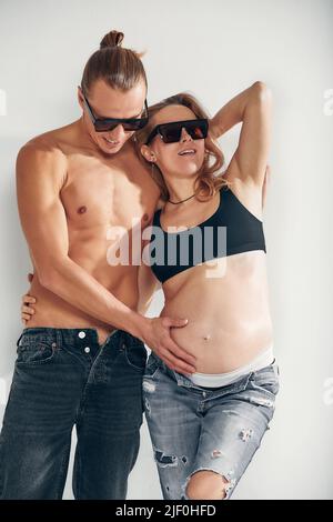 Gros plan d'un homme heureux embrassant le ventre de sa femme enceinte charmante debout dans la chambre blanche avec des lunettes de soleil. Papa attend la naissance du Banque D'Images