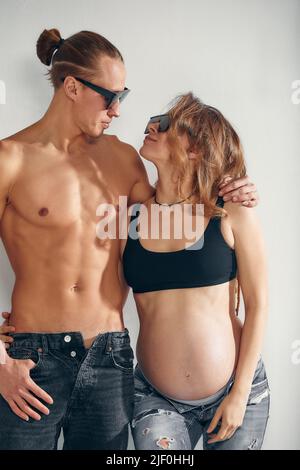 Gros plan d'un homme heureux embrassant le ventre de sa femme enceinte charmante debout dans la chambre blanche avec des lunettes de soleil. Papa attend la naissance du Banque D'Images