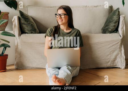 Jeune femme heureuse en lunettes travaille sur un ordinateur portable tout en étant assise sur le sol dans l'appartement. Banque D'Images