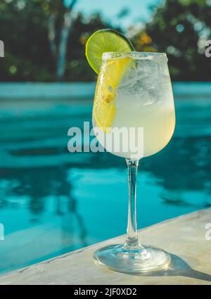 Cocktail glacé tropical au citron frais servi dans un verre transparent à côté de la piscine bleue avec reflet de l'eau à Tulum pendant l'heure d'or Banque D'Images