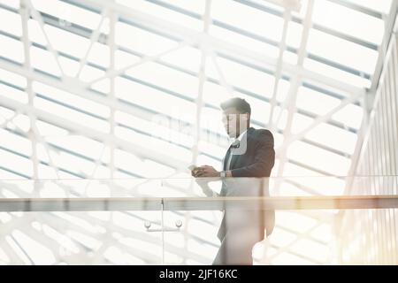 Un jeune homme d'affaires noir à la mode se penchait sur des rampes et envoyant des SMS tout en attendant un collègue dans le hall d'affaires Banque D'Images