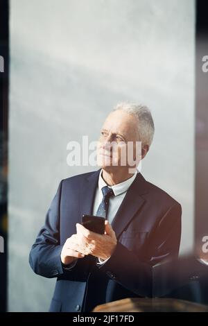 Le contenu est un homme d'affaires aux cheveux gris rêveux qui se tient en costume dans le lobby et utilise l'application gadget pour planifier le temps Banque D'Images