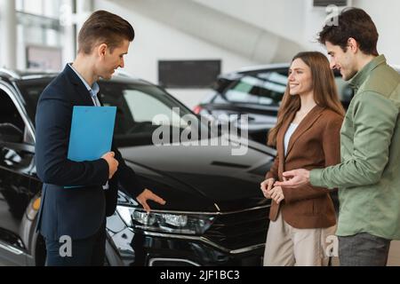 Jeunes conjoints positifs parlant au vendeur au sujet de l'achat d'une nouvelle voiture à l'atelier de l'établissement concessionnaire. Achat ou location de voiture Banque D'Images