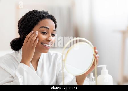 Belle jeune fille noire regardant dans le miroir et appliquant sous la crème des yeux Banque D'Images