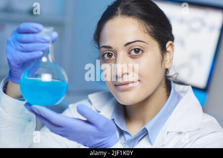 Portrait du contenu confiante jeune race mixte femme chimiste dans des gants de protection bleus tenant flacon de laboratoire de liquide cyan Banque D'Images