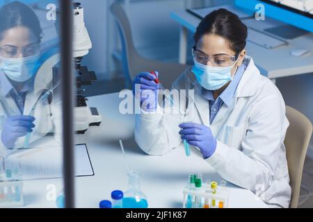 Jeune femme indienne sérieuse chercheuse chimique en vêtements de travail de protection assise au bureau en laboratoire et tombant du liquide bleu dans le tube à essai Banque D'Images