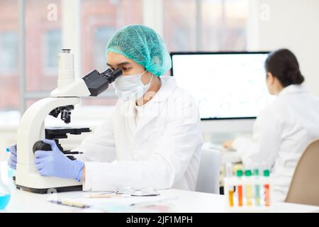 Jeune scientifique asiatique concentré et sérieux, en capuchon et masque stériles, assis à la table et analysant l'échantillon d'essai au microscope en laboratoire Banque D'Images