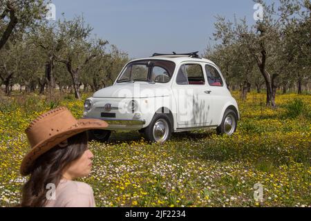 Image d'une femme assise au milieu d'un pré fleuri portant un chapeau de cow-boy avec une vieille voiture italienne d'époque en arrière-plan. Voyage en Toscane. Banque D'Images