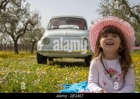 Adorable et belle petite fille souriante assise dans un pré fleuri avec une vieille voiture italienne Fiat 500 en arrière-plan. Vacances à Florence, Banque D'Images