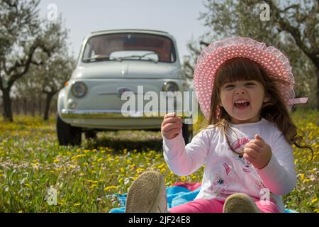 Image d'une petite fille gaie et souriante assise dans un pré fleuri avec une vieille voiture Fiat 500 vintage en arrière-plan. vacances en toscane Banque D'Images