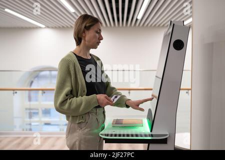 Femme d'âge moyen enregistrant un livre à la bibliothèque, en utilisant un terminal en libre-service Banque D'Images