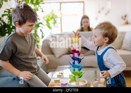 Petits frères et sœurs jouant avec le marbre en bois montessori dans la salle de séjour, leur mère est assise sur un canapé avec ordinateur portable. Banque D'Images