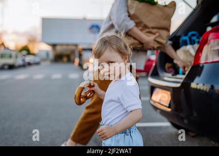 Une jeune mère avec une petite fille après avoir fait ses courses en tenant des sacs de shopping sans déchets avec une épicerie, en chargeant une voiture. Banque D'Images