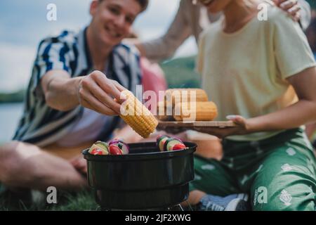 Gros plan de jeunes amis mettant du maïs sur le grill et ayant le barbecue lors du camping dans le terrain de camping. Banque D'Images