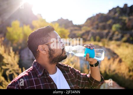 Un homme boit l'eau d'une bouteille de verre au soleil lors d'une randonnée Banque D'Images