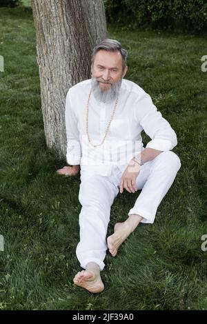 une longueur complète d'entraîneur de yoga barbu assis sur une pelouse verte et souriant à la caméra Banque D'Images